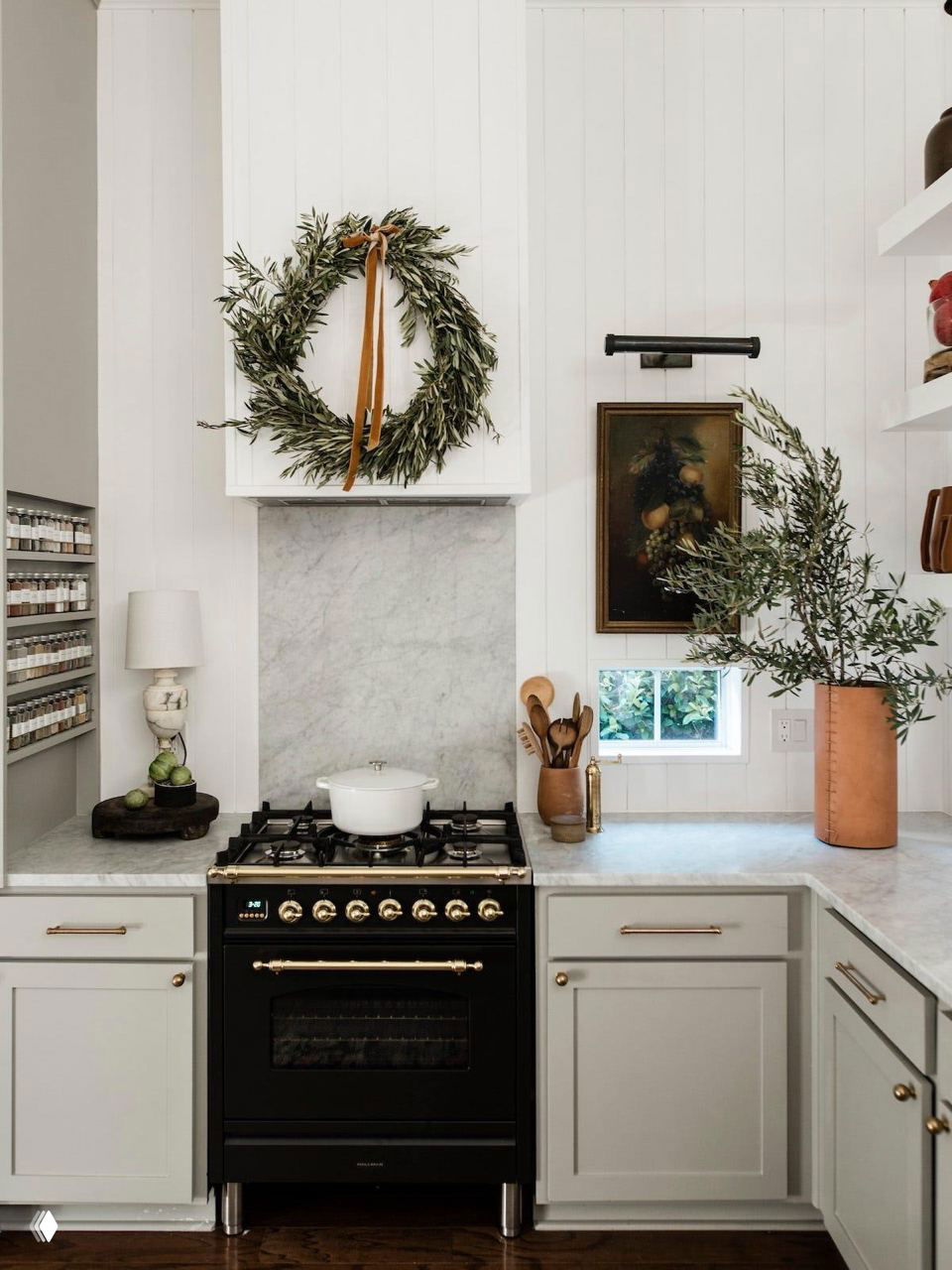 Небольшая кухня со чёрной плитой, венком над вытяжкой и зелёными растениями на столешнице — классическая рождественская декорация в деревенском стиле.