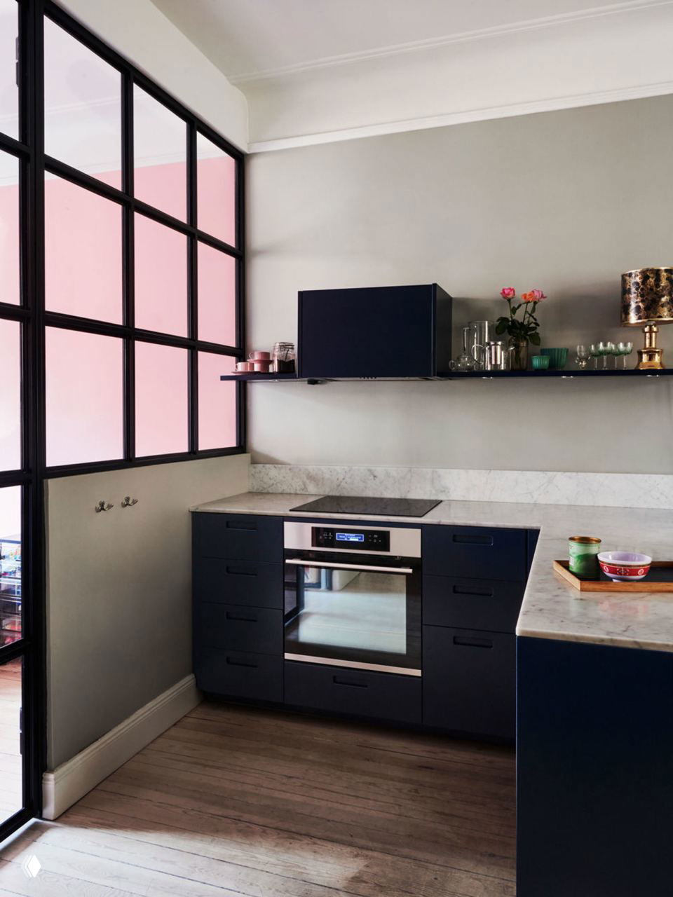 Кухонный уголок в небольшой квартире: тёмно‑синие фасады, мраморная столешница, стеклянная перегородка и дневное естественное освещение.