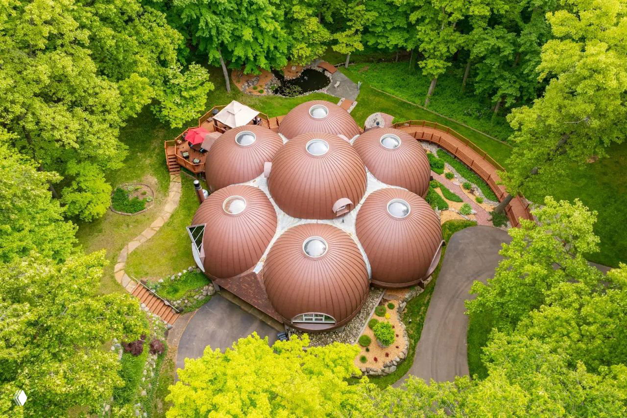 Аэрофотоснимок «Лукового дома» среди густого леса округа Каламазу — семь конических куполов с круглыми иллюминаторами и подъездная дорожка.