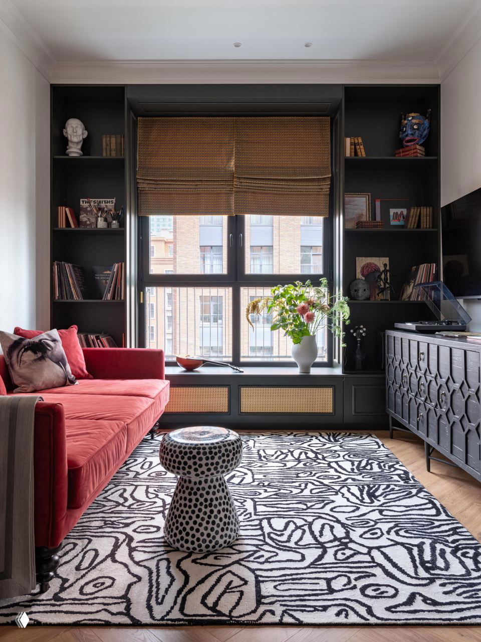 Гостиная в московской квартире: красный бархатный диван, чёрные встроенные стеллажи вокруг окна, графичный ковёр и украшения на полках.