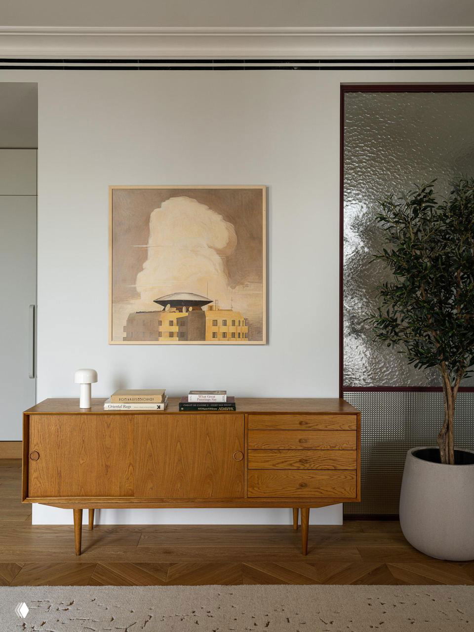 Винтажная низкая деревянная тумба с книгами и небольшой скульптурой, над ней пастельная картина и текстурное остекление в сдержанном интерьере