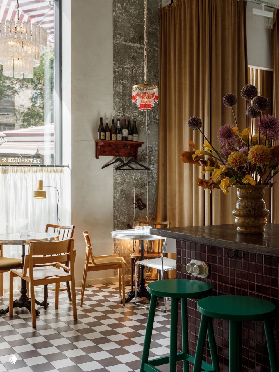 Интерьер бистро с зелёными барными стульями и стойкой, шахматной плиткой на полу, высоким окном с занавеской и вазой с цветами.