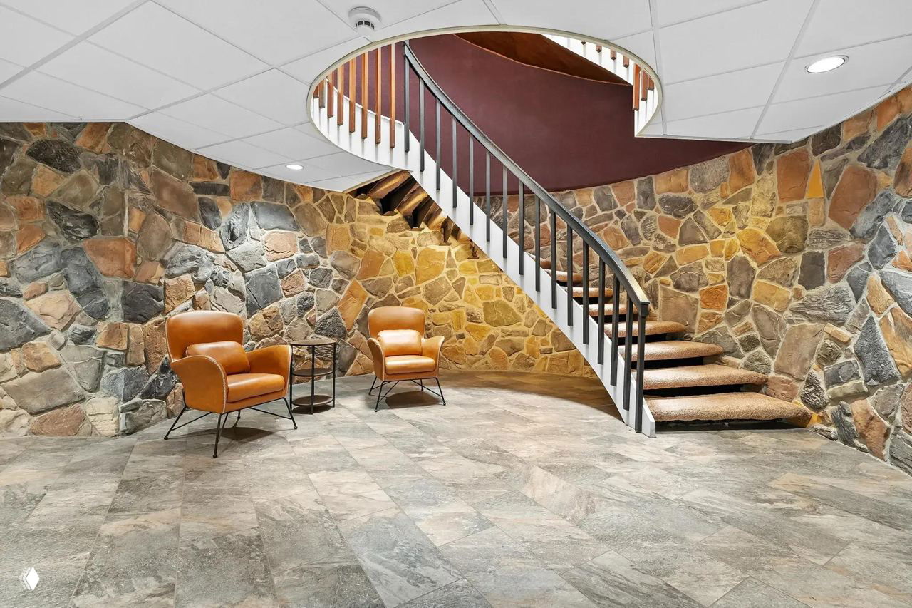 Интерьер цокольного этажа с винтовой лестницей и стеной из природного камня — зона отдыха с двумя кожаными креслами и аккуратным полом.