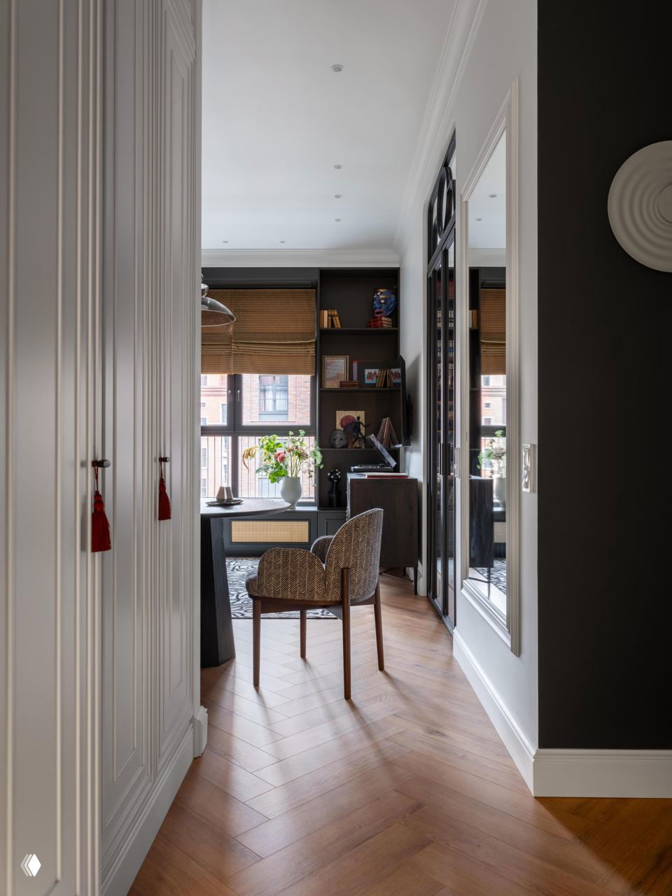 Вид из коридора на рабочую зону у окна: стул у окна, вазон с цветами, встроенные шкафы и отражение пространства в зеркале.