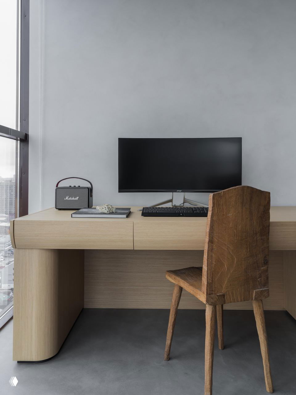 Рабочая зона у окна: деревянный стол с закруглёнными углами, монитор и стул из состаренного дерева, вид на городской пейзаж за панорамным окном.