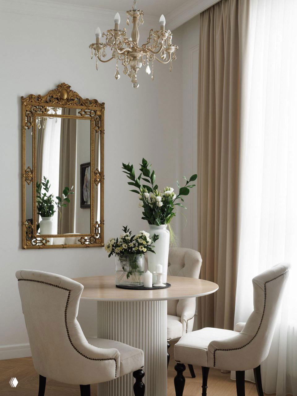 Обеденная зона: круглый деревянный стол, стулья с обивкой светлого тона, хрустальная люстра и большое золочёное зеркало на стене, создающие атмосферу французской классики.