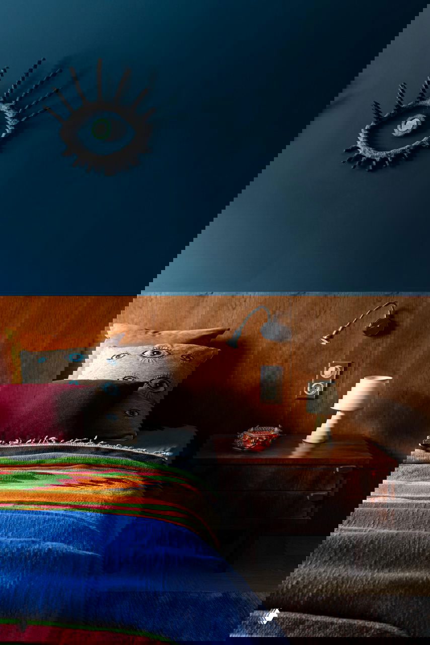 Спальня с темно-синей стеной и деревянным изголовьем, цветные подушки и покрывало, декоративное арт-украшение в виде глаза над кроватью.