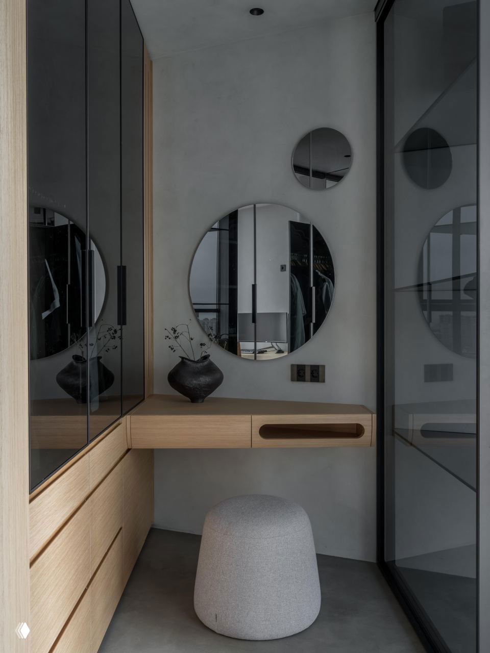 Гримерная зона с круглым зеркалом, деревянными фасадами и мягким пуфом в минималистичном светло-сером интерьере, отражение пространства в стекле.