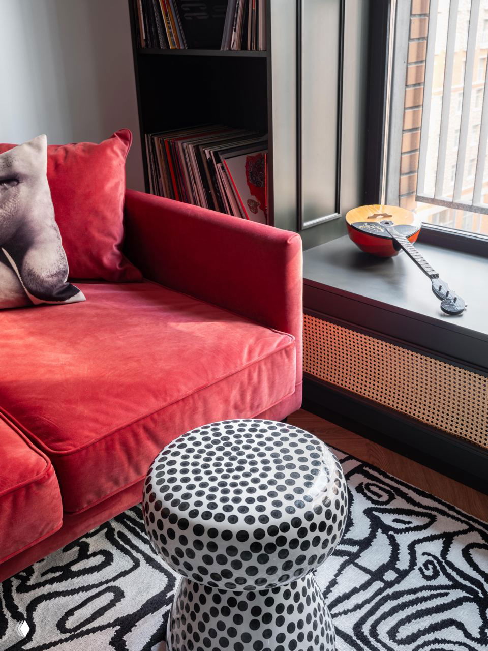 Крупный план на зону отдыха: бархатный красный диван, декоративный пуфик в горох и подоконник с музыкальным инструментом и коллекцией винила.