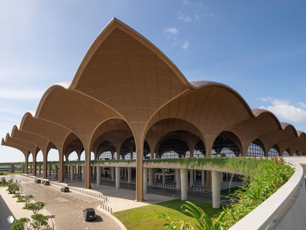 Внешний вид терминала аэропорта Течо в Пномпене: волнообразная стальная крыша с решетчатым узором и рядами колонн под ней, дневной пейзаж.