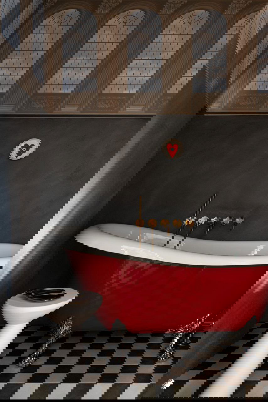 Ванная комната с красной чугунной ванной на ножках, черно-белой шахматной плиткой на полу и сдержанными серыми стенами с декоративным фризом у потолка.