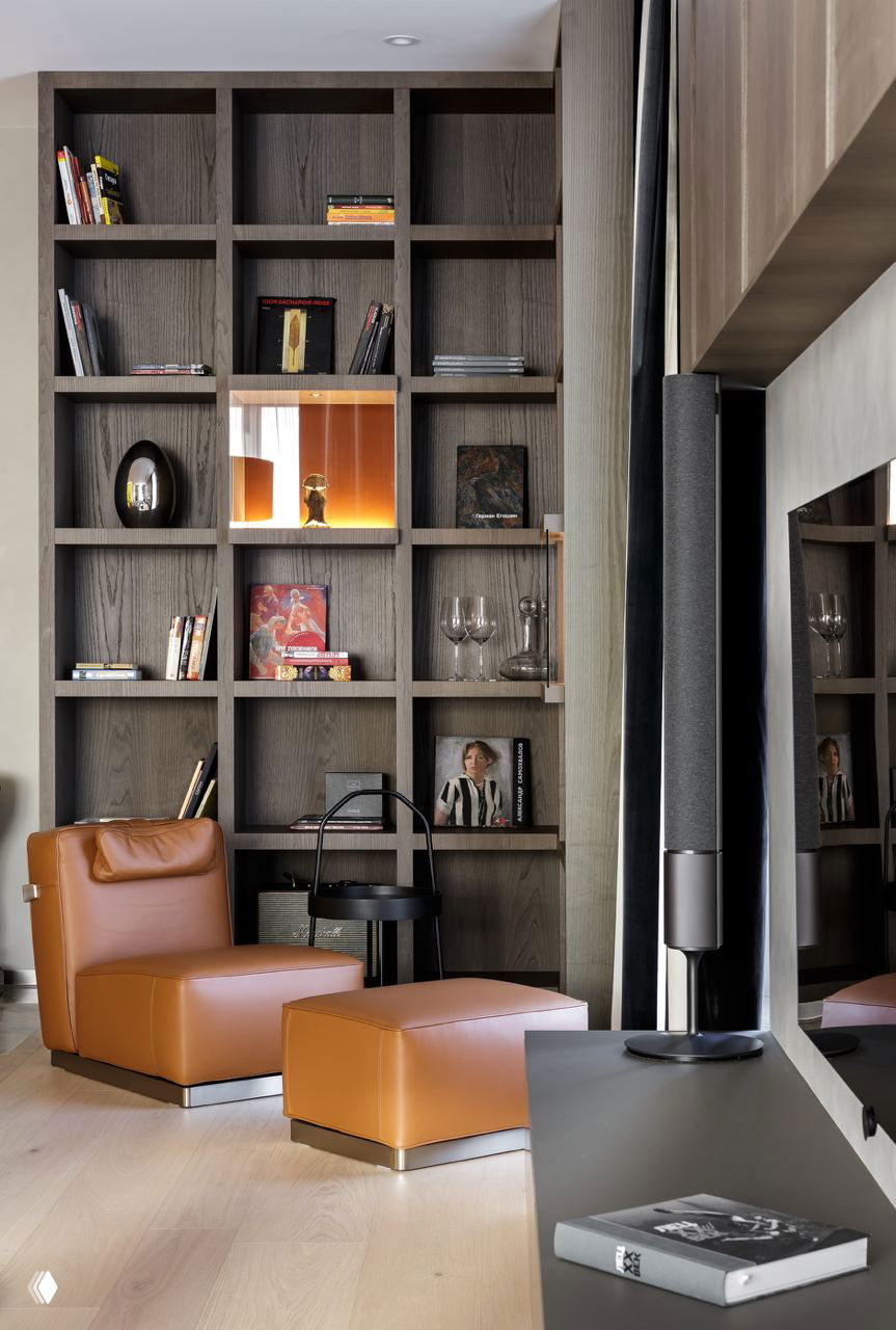 Угол гостиной с модульной деревянной полкой-решёткой, аксессуарами, книгами и коньячным кожаным креслом — акцент на текстурах и хранении коллекции.