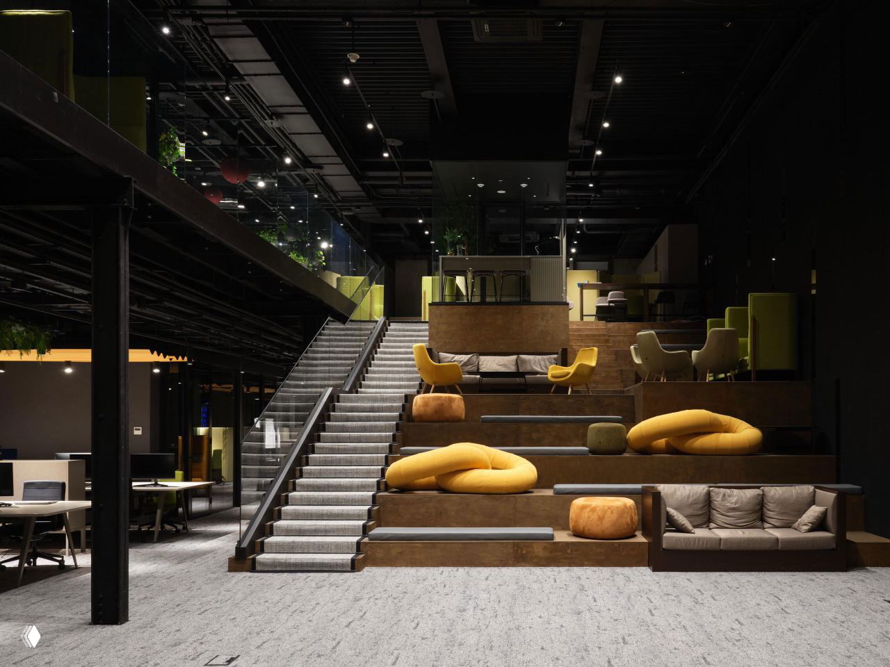Вид внутри двухсветного амфитеатра офиса: ступени-места для сидения, широкая лестница и крупные жёлтые мягкие пуфы на фоне чёрного потолка и подсветки.