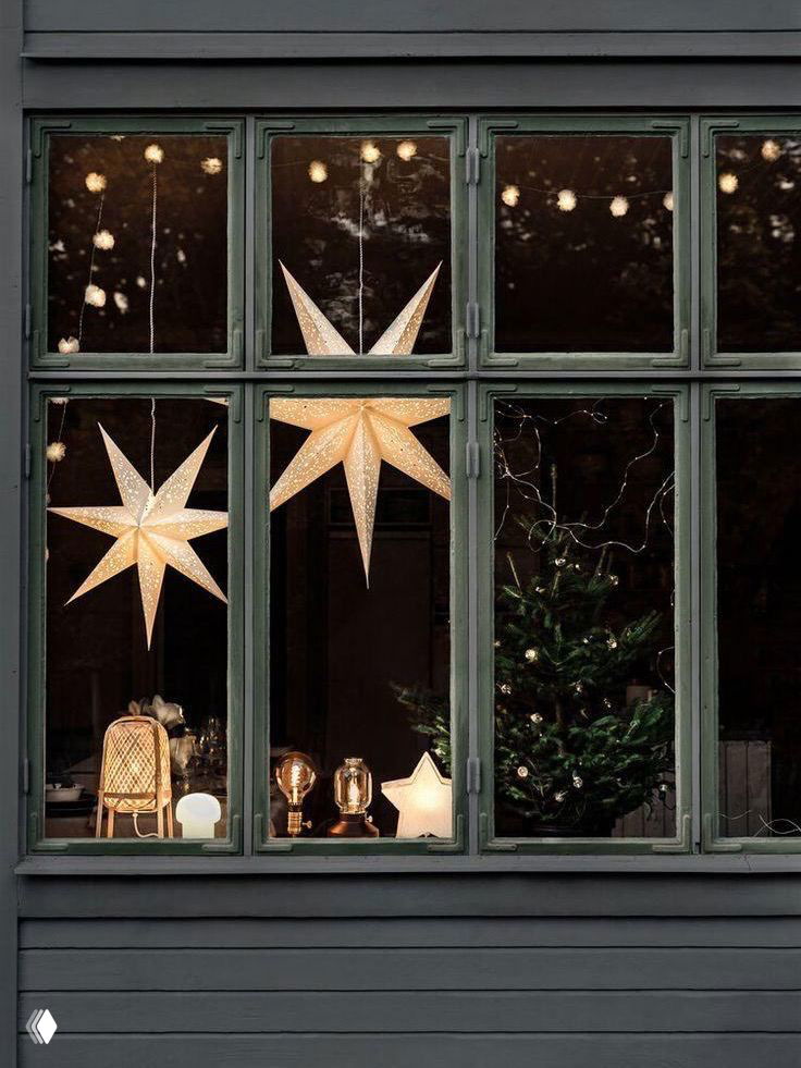 Окно с висящими бумажными звёздами и тёплой подсветкой: тёмный наружный фон и интерьерная лампа создают уютный вечерний праздничный антураж.