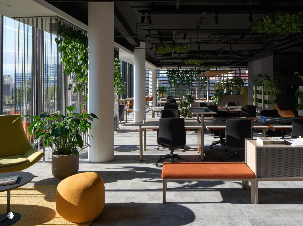 Открытая рабочая зона с рядами столов у панорамных окон, естественное освещение, зелёные растения и разноцветные мягкие сиденья и пуфы.