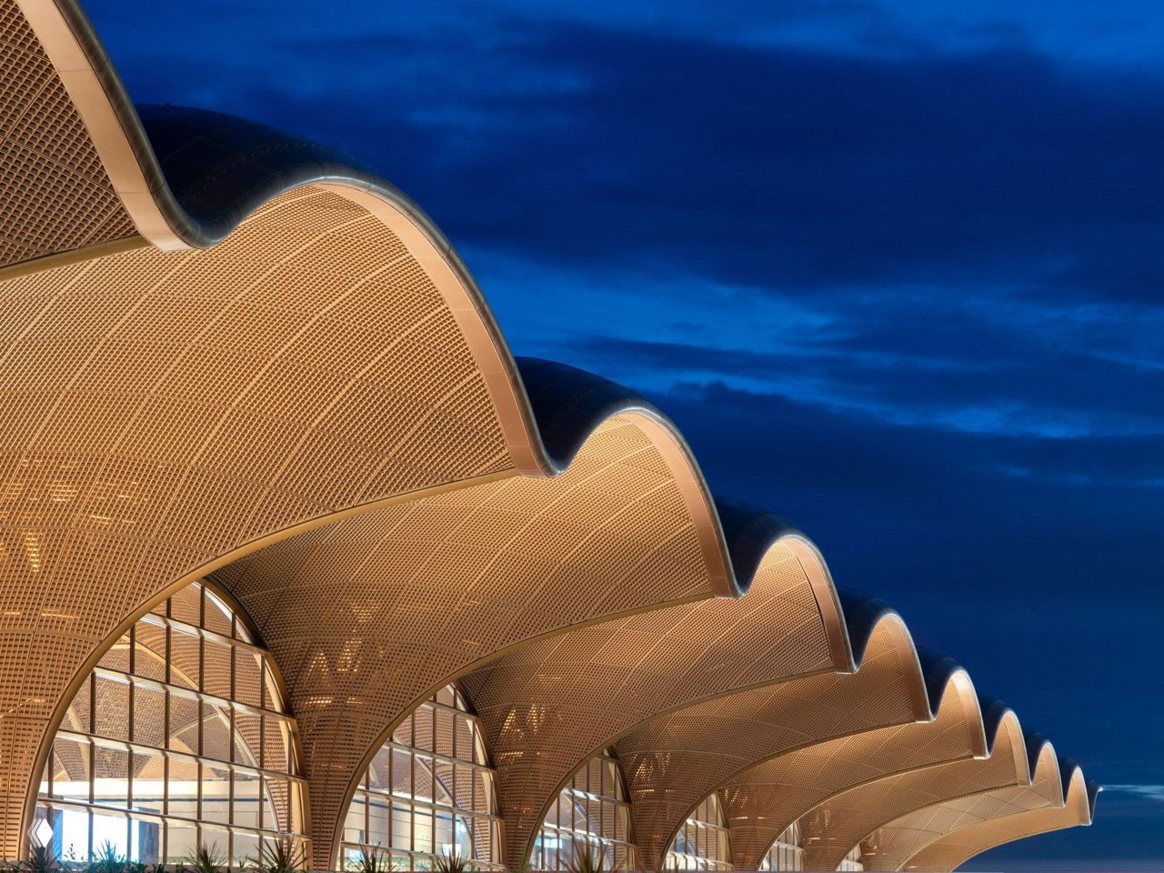 Фрагмент крыши и фасада при вечерней подсветке: изогнутые арочные модули, решетчатая текстура панелей и глубокое синее небо на заднем плане.