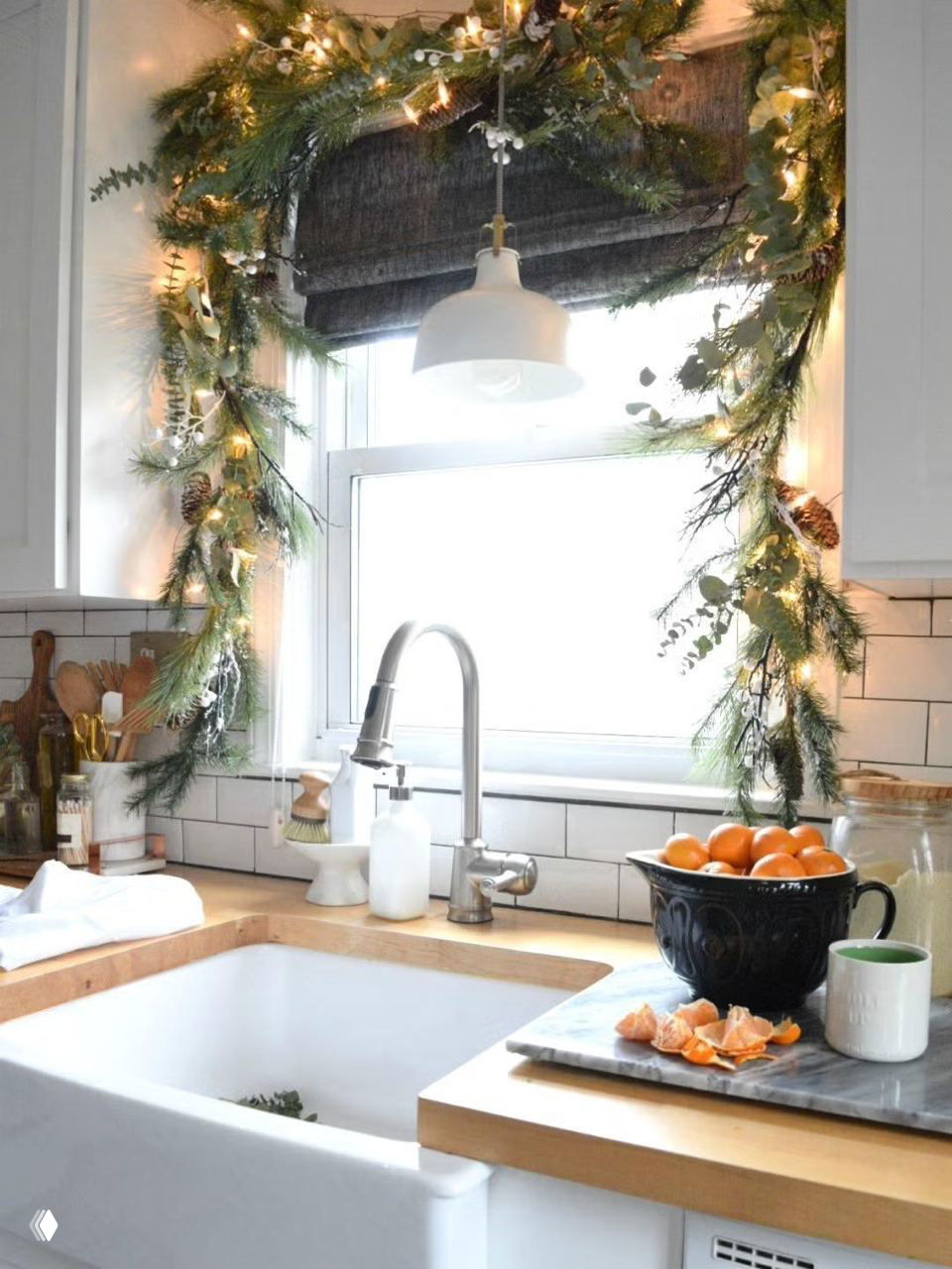 Кухонное окно, украшенное хвойной гирляндой с огоньками; на столе миска с мандаринами и простой декор для уютной праздничной атмосферы.