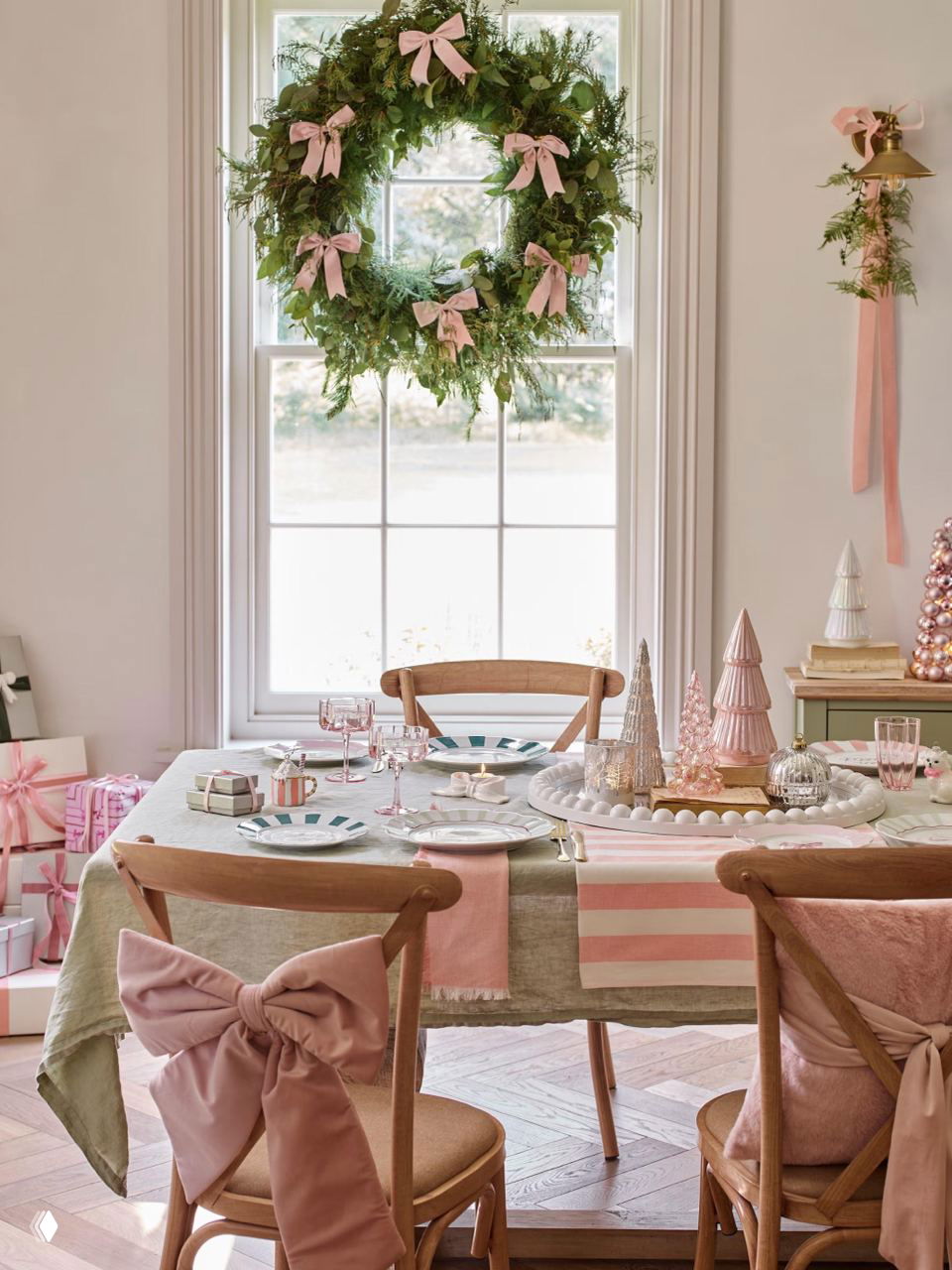 Большой живой венок с розовыми лентами, размещённый в проёме окна над сервированным столом в пастельной гамме — нежная праздничная композиция.