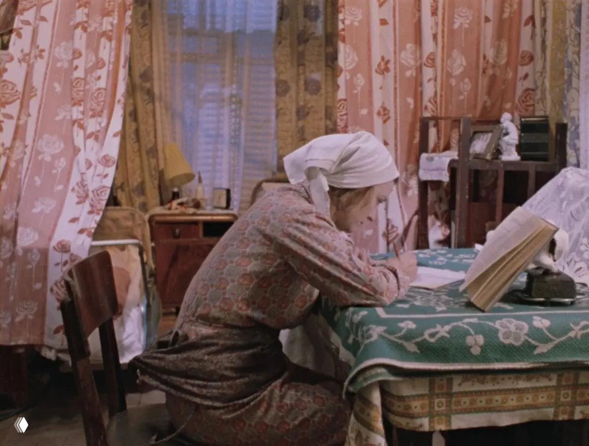 Интерьер комнаты с цветными занавесками и столом, где женщина в платке пишет за столом; вокруг видны предметы быта и кровать.