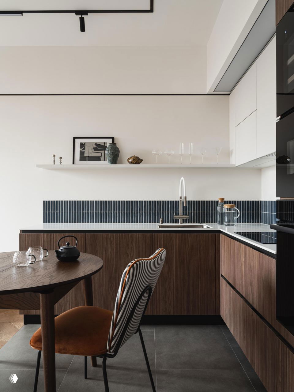 Кухня с тёмной столешницей и деревянными фасадами, узкая декоративная плитка‑фартук вдоль стены и обеденный стол с элегантным стулом на фоне простой белой стены.