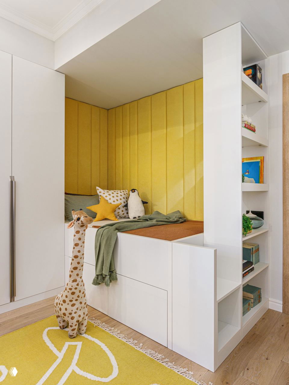 Уютная детская зона со встроенным спальным местом, яркой жёлтой мягкой панелью, игровым ковриком и открытыми полками для книг и игрушек рядом со спальным местом.