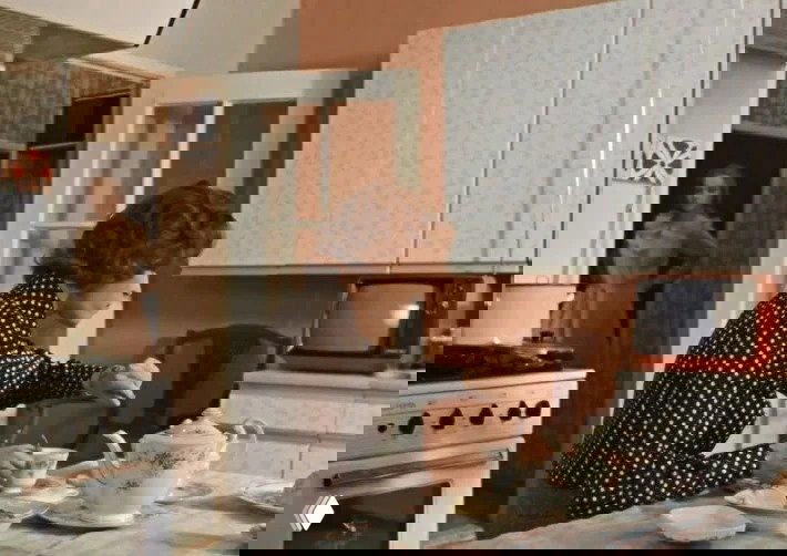 Кухонная сцена: плита, сервиз на столе и женщина, сидящая за столом — бытовая зона в квартире героини с характерной посудой.