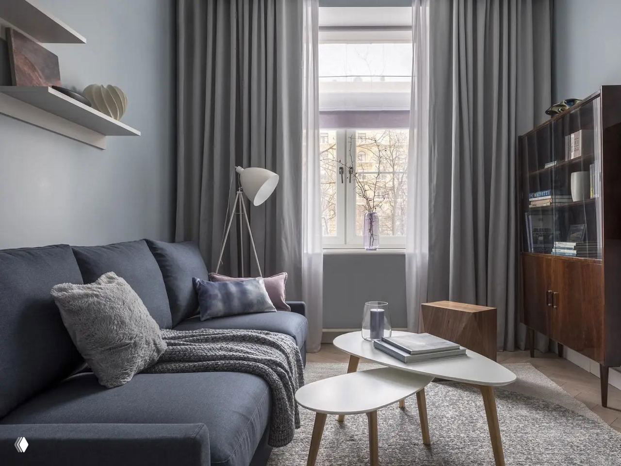 Гостиная с тёмно‑синим диваном‑кроватью на переднем плане, кофейные столики и большое окно позади — интерьер в спокойных тонах.