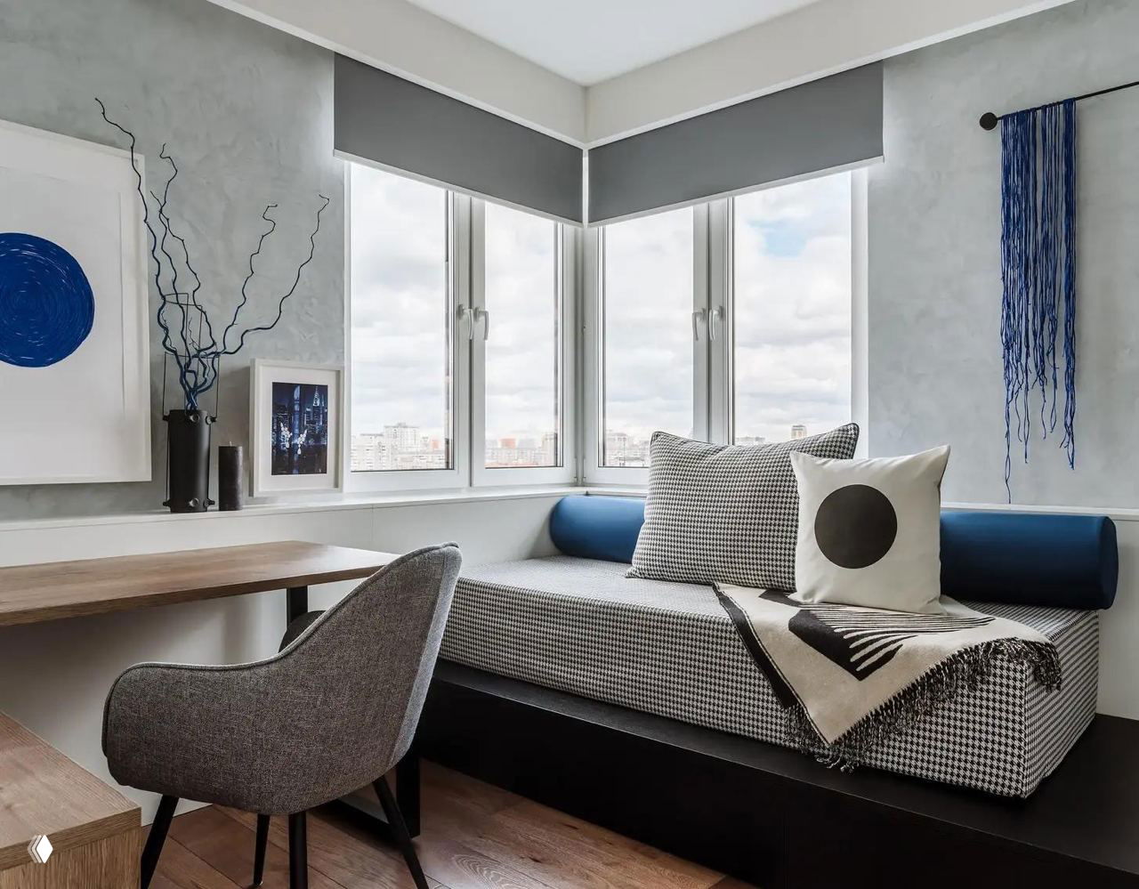 Кушетка/диван‑кровать у окна в нише с деревянным рабочим столом рядом, клетчатая обивка и декоративная подушка на фоне города за окном.