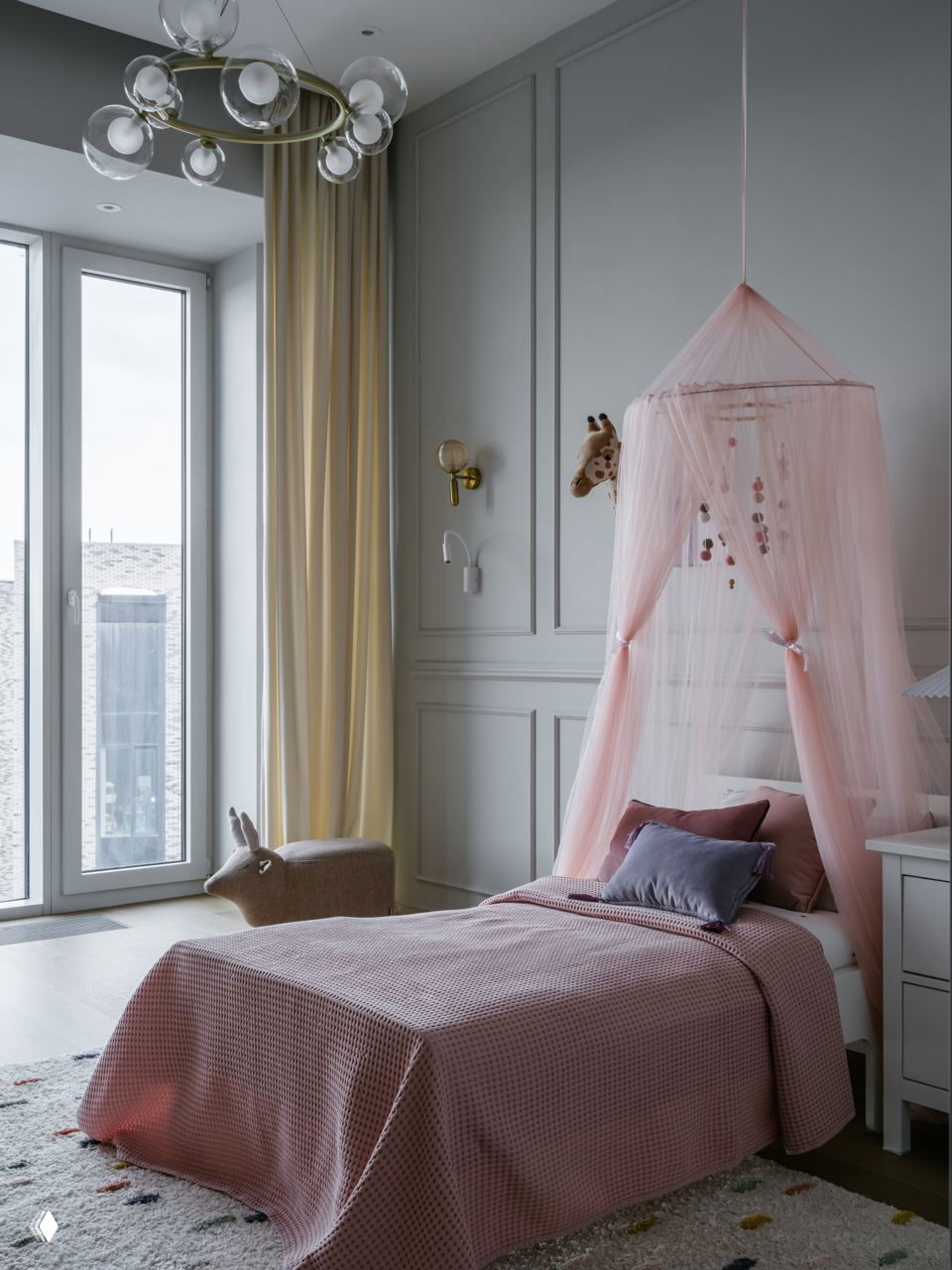 Серая элегантная спальня ребёнка с высоким окном, розовой тюлевой накидкой-надстройкой над кроватью и сдержанными текстильными решениями в пастельной палитре.