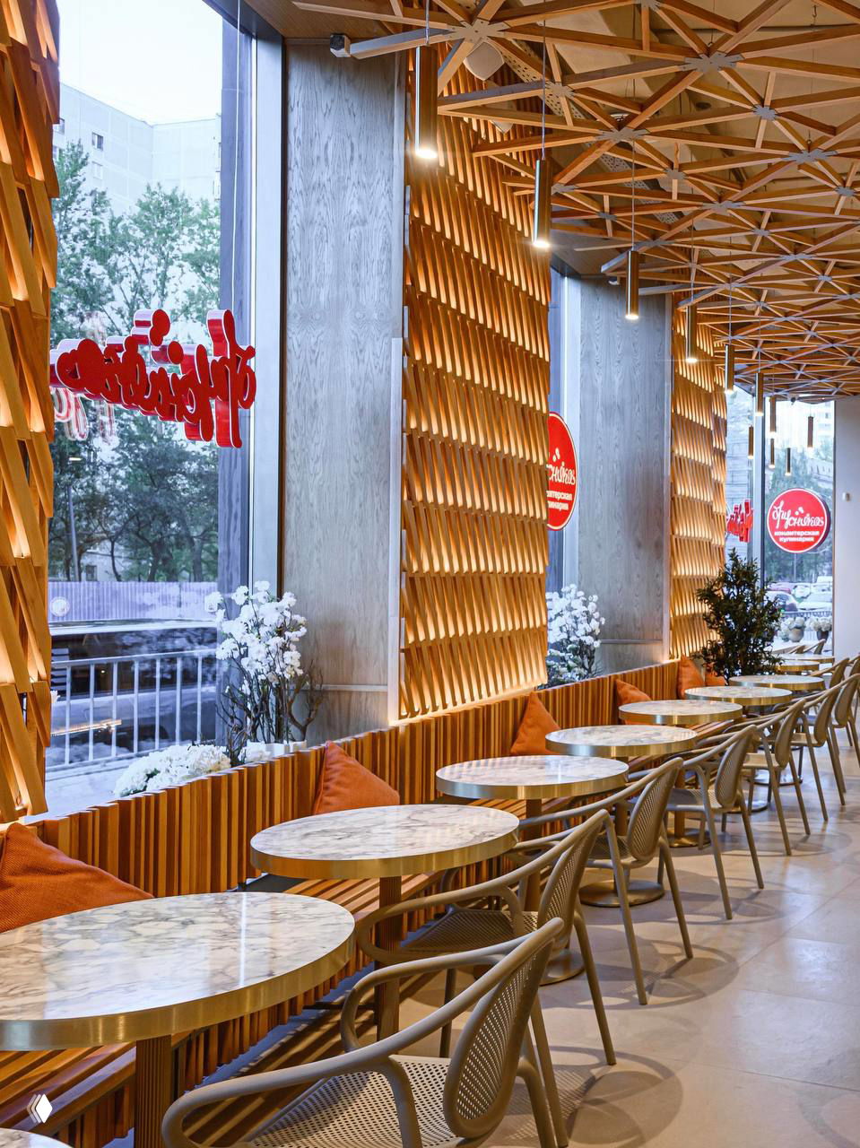 Ряд столов вдоль окна, декоративные подушки, деревянные панели и геометрический потолок с диммируемыми светильниками создают тёплый интерьер.