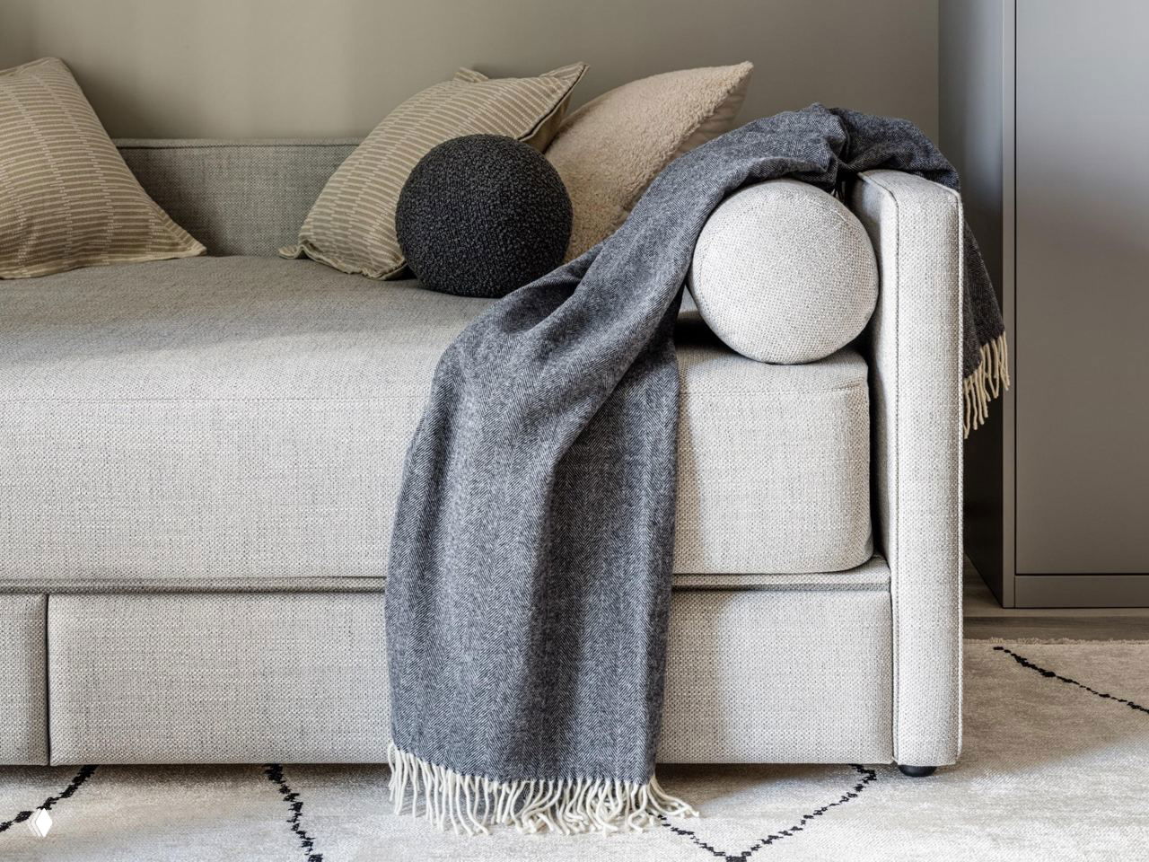 Фрагмент светлого дивана‑кровати с декоративными валиком и пледом, акцент на фактуре ткани и уютной текстиле на полу.