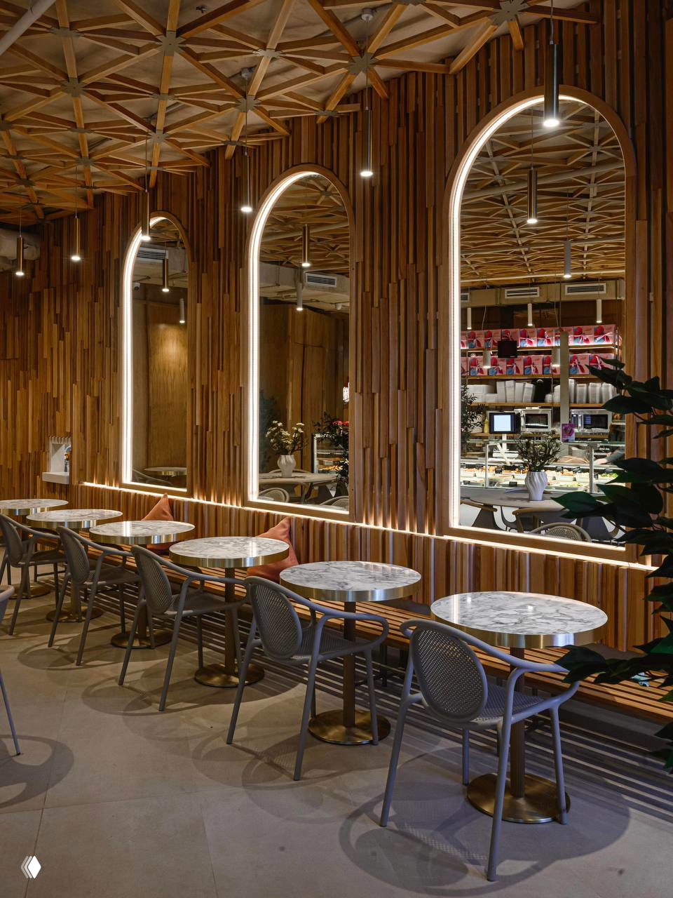 Арочные зеркала в деревянной обшивке, мраморные столешницы и выразительный деревянный потолок — акцент на текстуре и свете в интерьере кафе.
