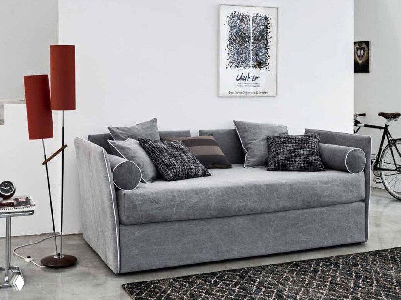 Серый прямой диван‑кровать с несколькими декоративными подушками и торшером рядом, стильный городской интерьер с ковром внизу.