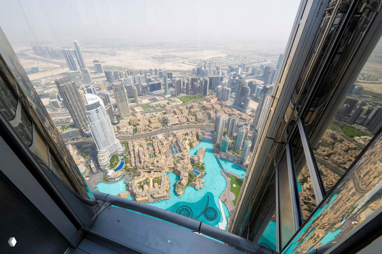 Вид с очень высокого этажа вниз на плотную застройку, стеклянные фасады небоскрёбов и ярко-голубые бассейны; сильное ощущение высоты и перспективы.