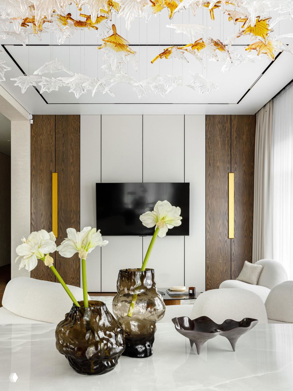 Обеденная/гостевая зона с декоративной стеклянной люстрой, тёмными деревянными панелями и прозрачными вязанными вазами на столе в тёплой палитре.