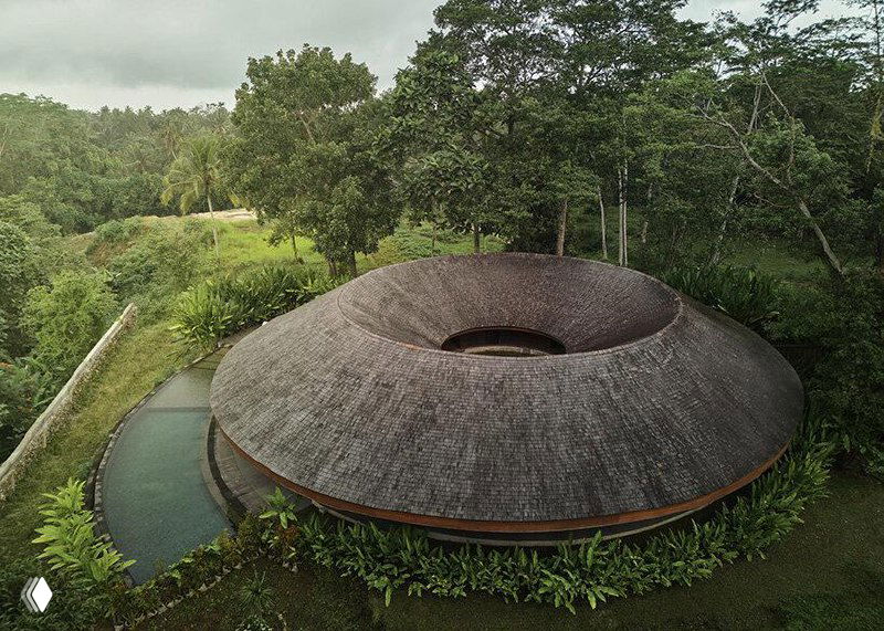 Аэрофотоснимок виллы-кольца на склоне под Убудом: круглая кровля из деревянного гонта и встроенный внутренний двор на фоне тропического леса.