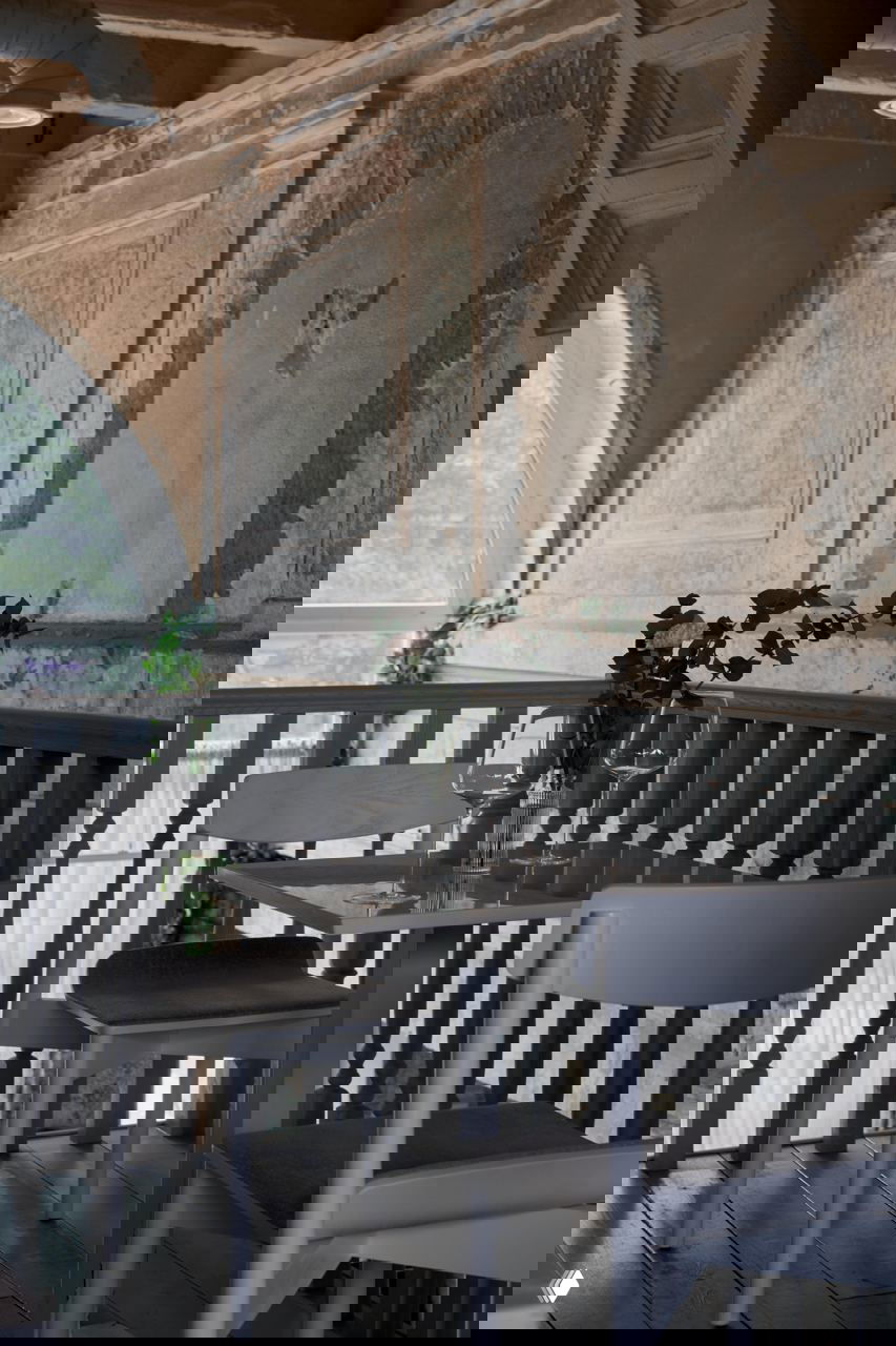 Уютный небольшой столик у перил второго уровня на фоне потертой декоративной штукатурки и арочного окна — сочетание старых стен и светлой мебели.