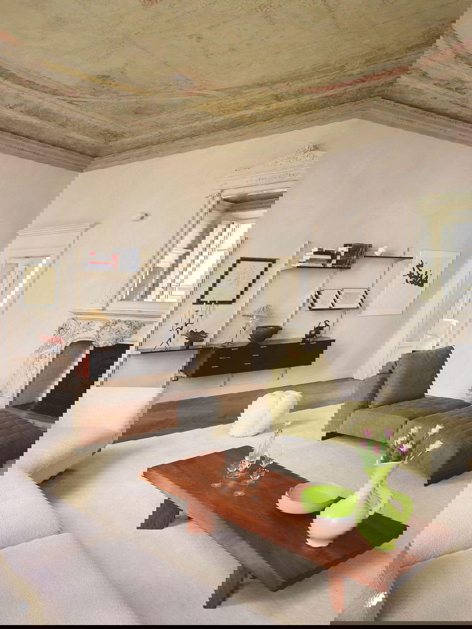 Просторная гостиная в светлых тонах с низким деревянным журнальным столиком, зелёным шезлонгом и пастельным декором на фоне фрески.