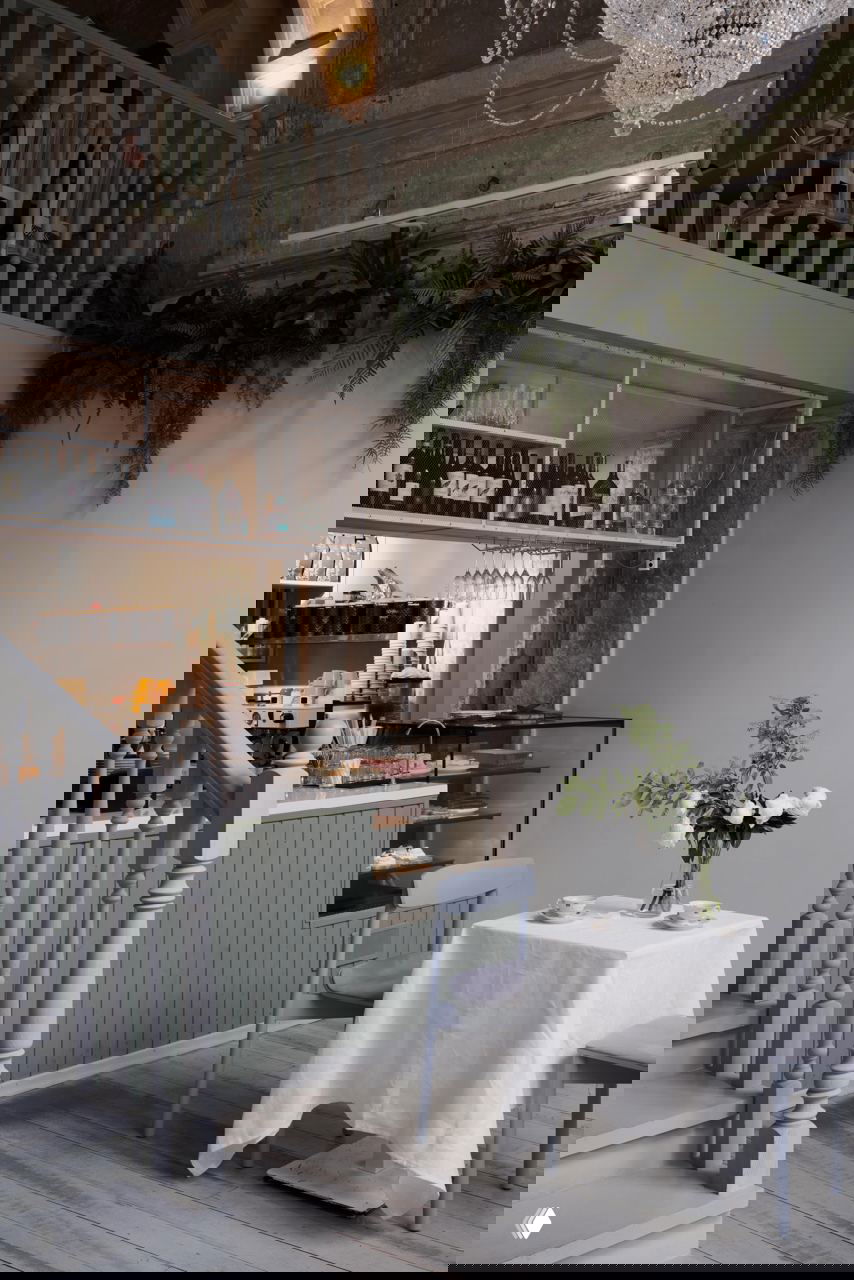 Барная зона и столик в светлых скандинавских тонах: пастельно‑серый кофейный прилавок, выставленные напитки и сервировка на фоне лестницы и люстры.