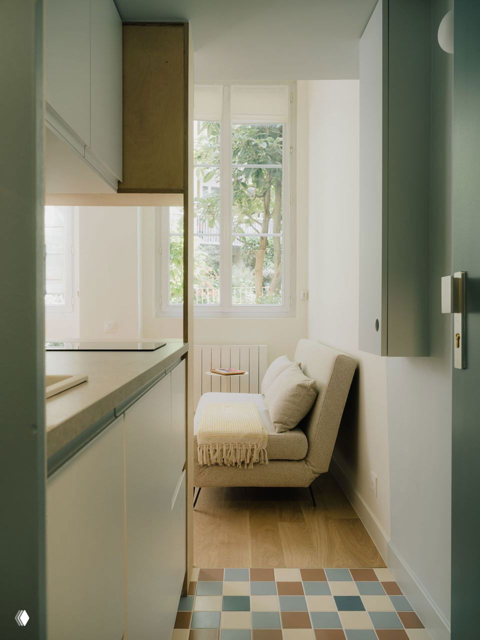 Вид через кухонный проход на компактную гостиную студии: небольшой диван у окна, светлый паркет и разноцветная мозаика в проходной зоне, создающие уютную атмосферу.