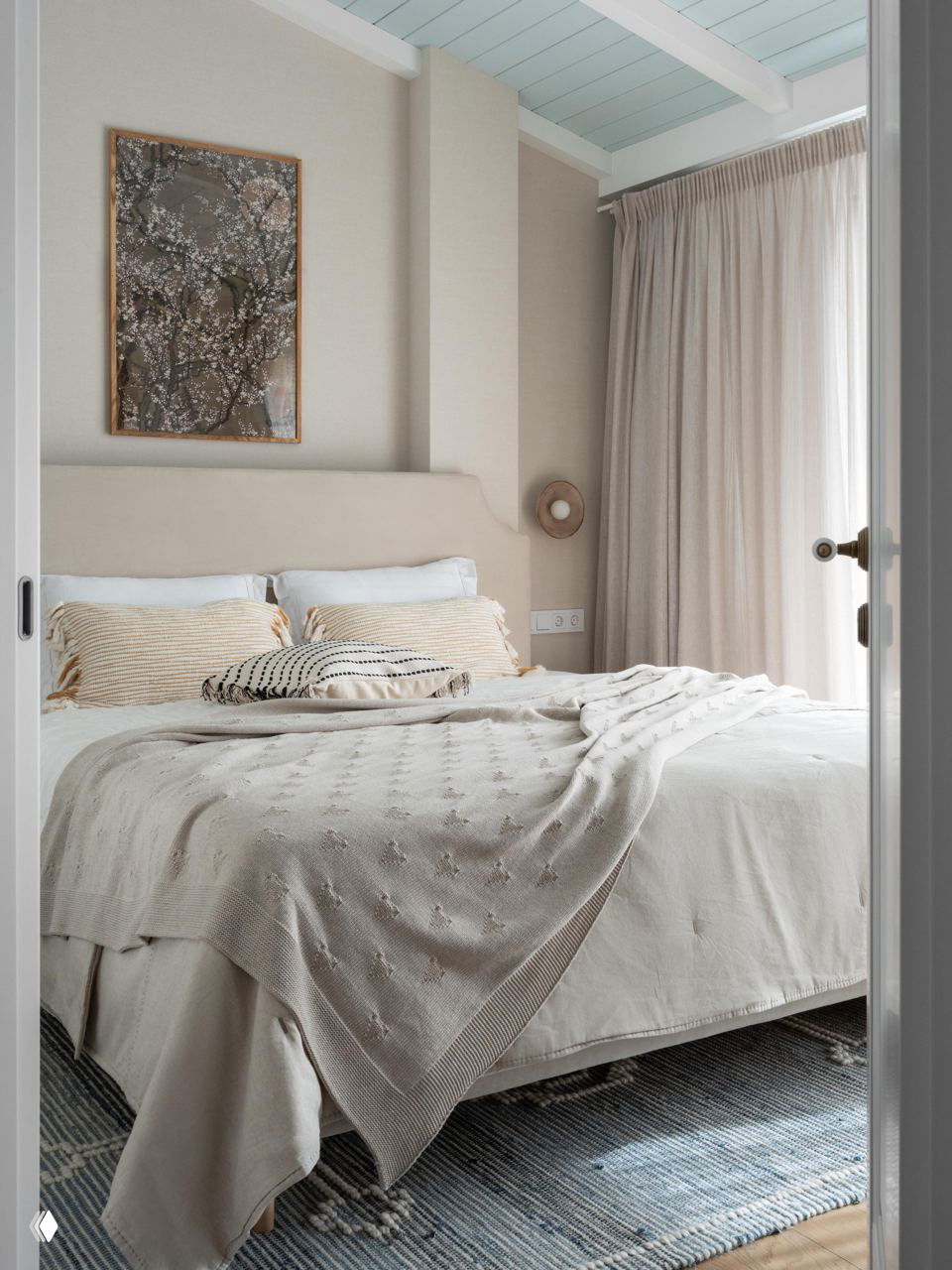Уютная спальня в нейтральных бежевых тонах: кровать с текстильным покрывалом, светлые шторы и мягкий ковёр у изголовья — спокойная атмосфера.