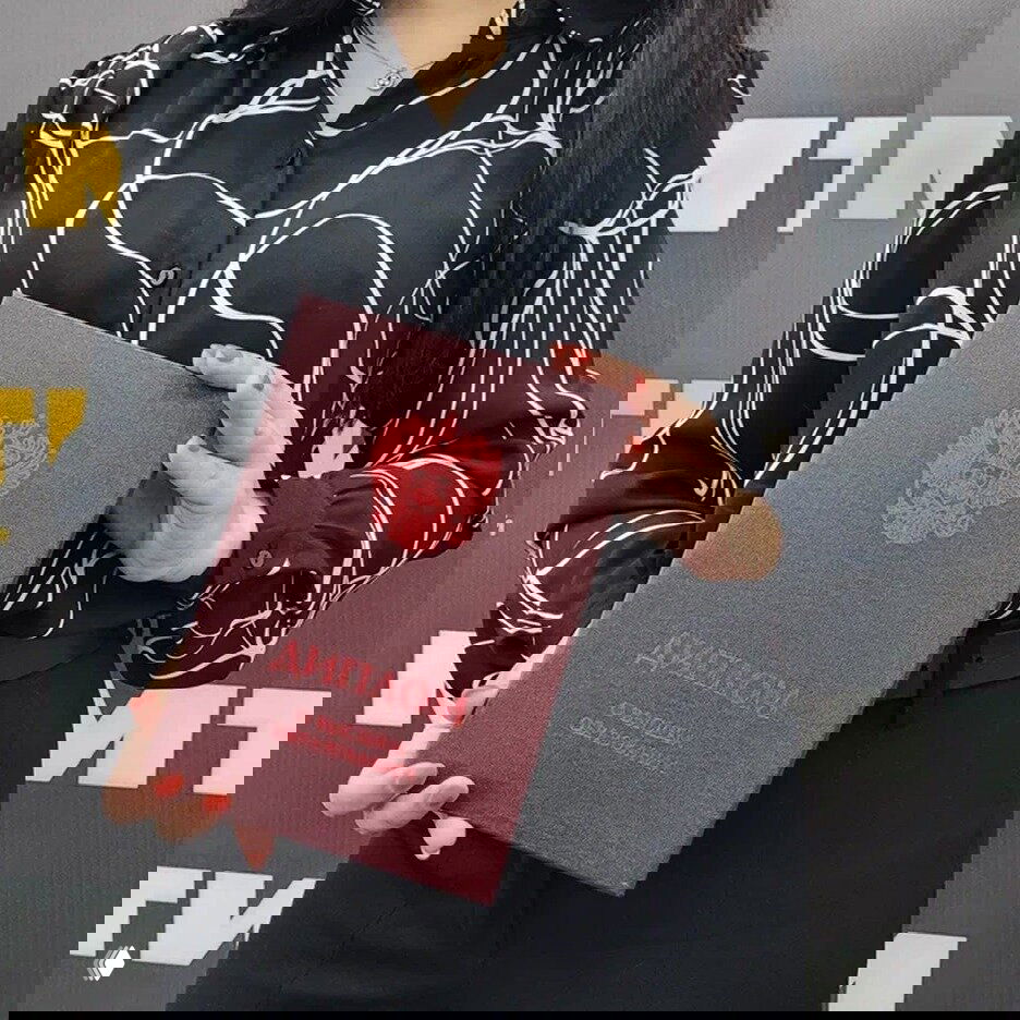 Женщина в деловой одежде держит бордовый переплёт с гербом РФ и надписью «Диплом о высшем образовании», на фоне фирменного стенда офиса.