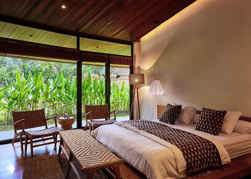 Интерьерная фотография спальни с деревянным потолком и большими проёмами, где видна связь внутреннего пространства с зеленью за пределами.