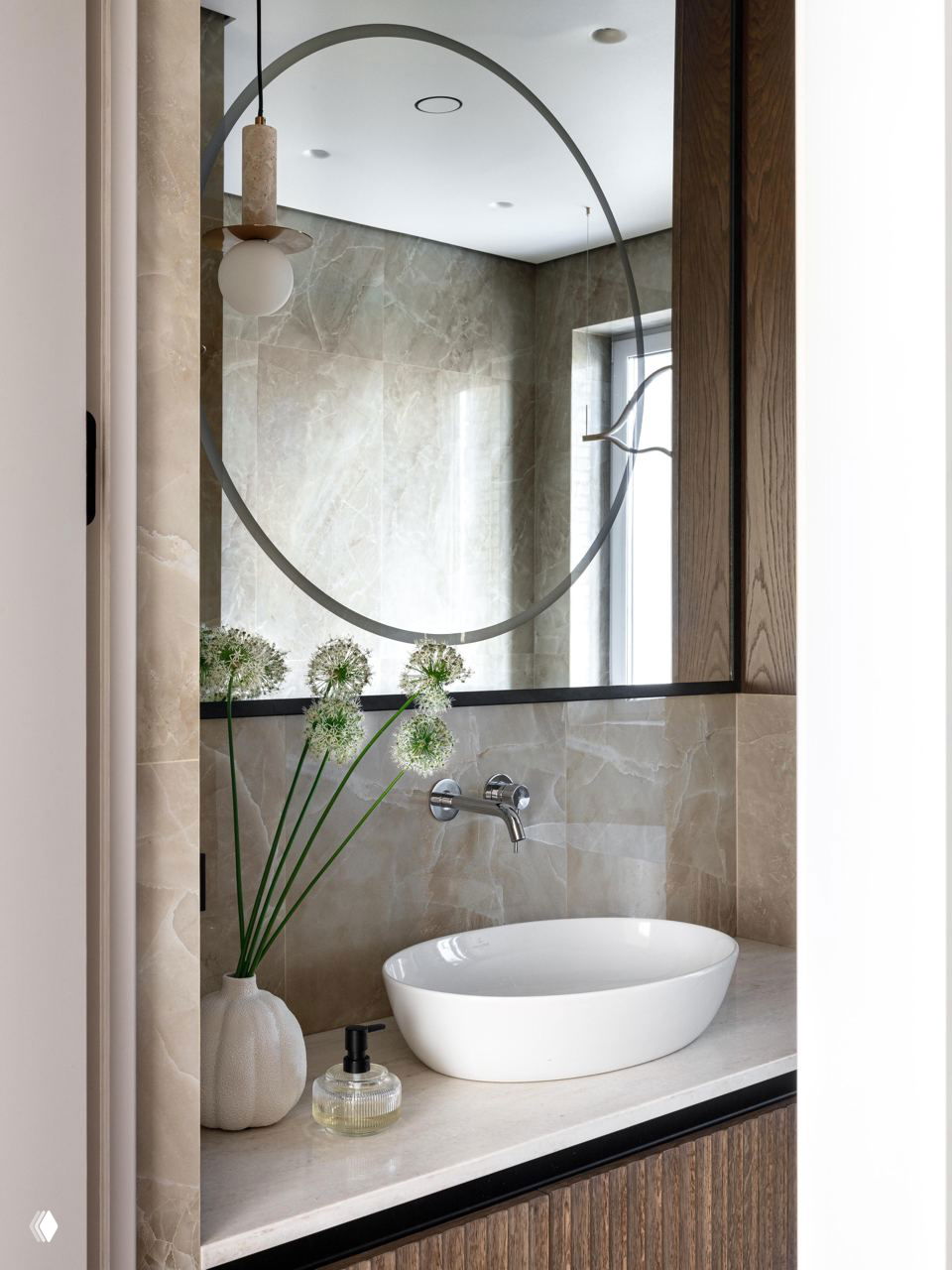 Ванная в тёплых бежевых тонах с овальной белой раковиной, круглым зеркалом и аккуратными минималистичными элементами декора на каменной столешнице.