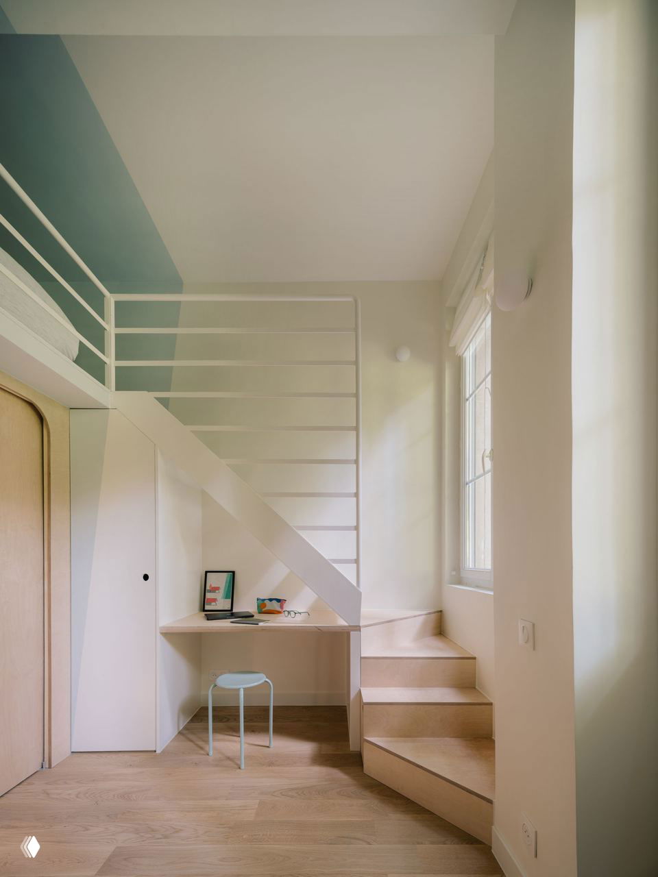Рабочая зона под лестницей: встроенный стол, компактный табурет и большие окна, впускающие естественный свет и визуально расширяющие маленькую студию.