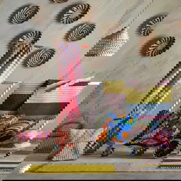 Сцена с ковриками, подушками и текстилем в зелёно‑жёлтой и розовой палитре; на потолке виден плетёный светильник, акцент — тканые принты.