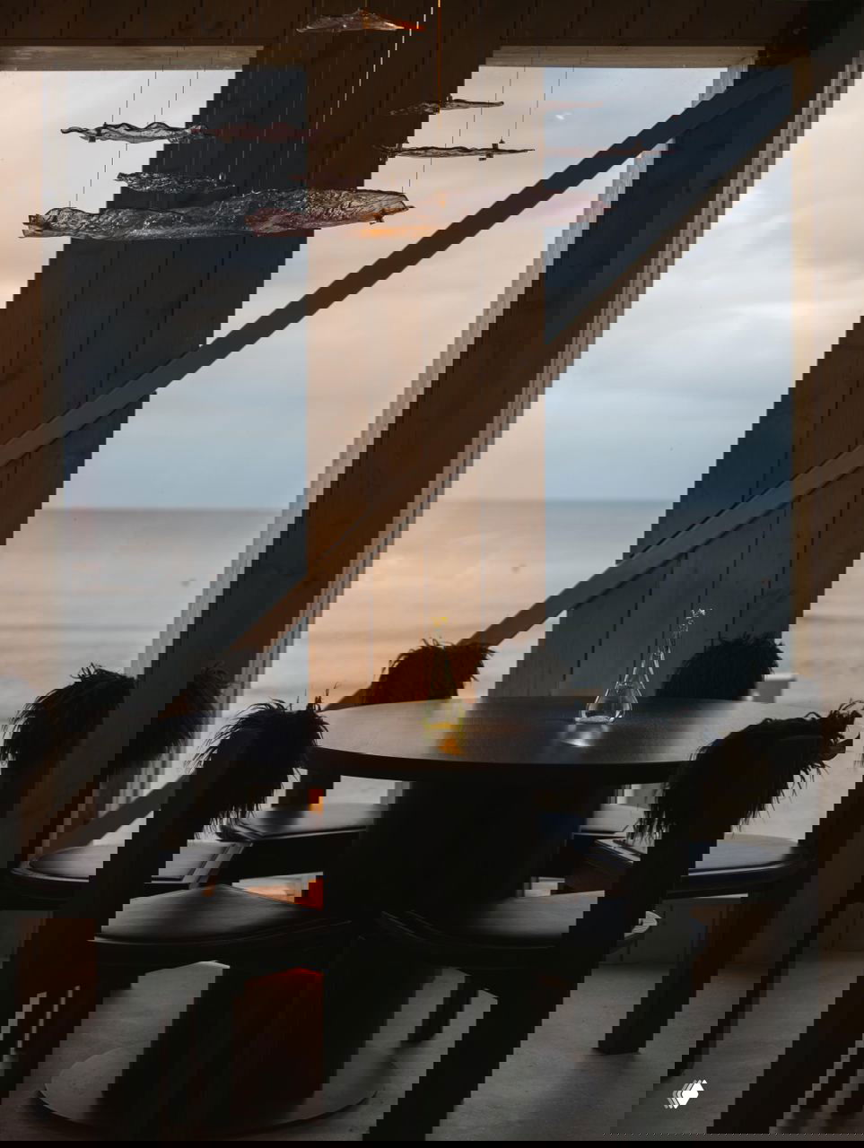 Круглый стол у окна с видом на Балтийское море: чёрные стулья с меховыми спинками, стеклянная бутылка на столе и приглушённая морская палитра в интерьере.