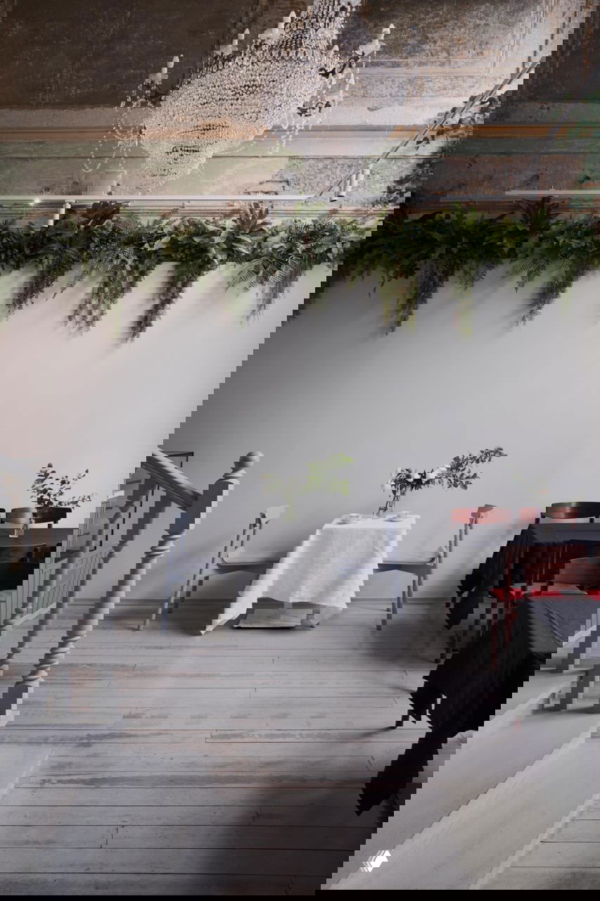 Минималистичный стол с белой скатертью в просторном зале, над ним люстра и зелёные подвесные растения — контраст простоты и декоративности в интерьере.