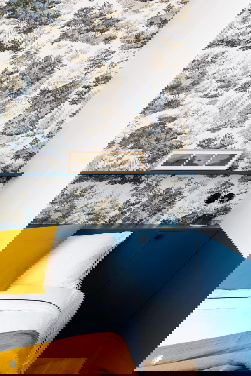 Детали спальни: декоративные пейзажные обои, тёмно‑синяя покраска нижней части стены и яркие жёлтые текстильные акценты на кровати в небольшой комнате.