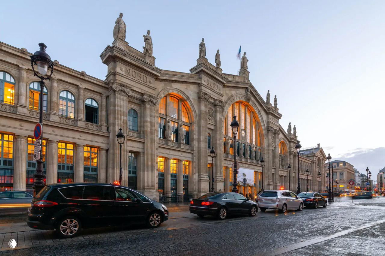 Фасад исторического железнодорожного вокзала в вечернем освещении: большие арочные окна, скульптуры на карнизе и тёплый свет внутри